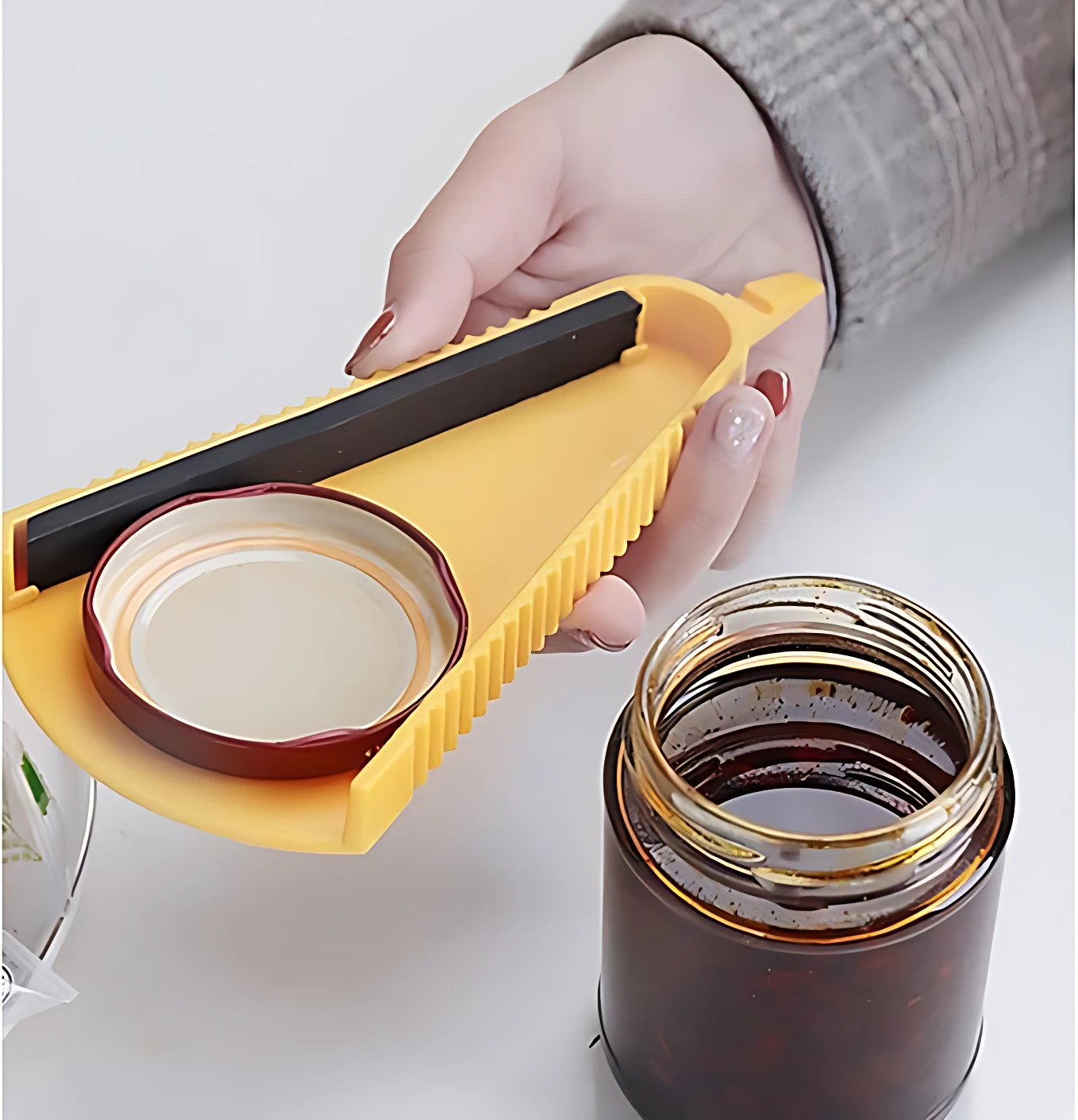 Abridor de botellas palanca antideslizante en amarillo, con mango negro y soporte para tapa de vidrio. Se muestra en uso sobre frasco de vidrio con contenido oscuro.