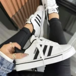 Zapatilla Adidas Campus blanca con tres rayas negras, suela acolchada y cordones blancos, vista desde arriba con pie derecho levantado y mano sosteniendo el lateral izquierdo.