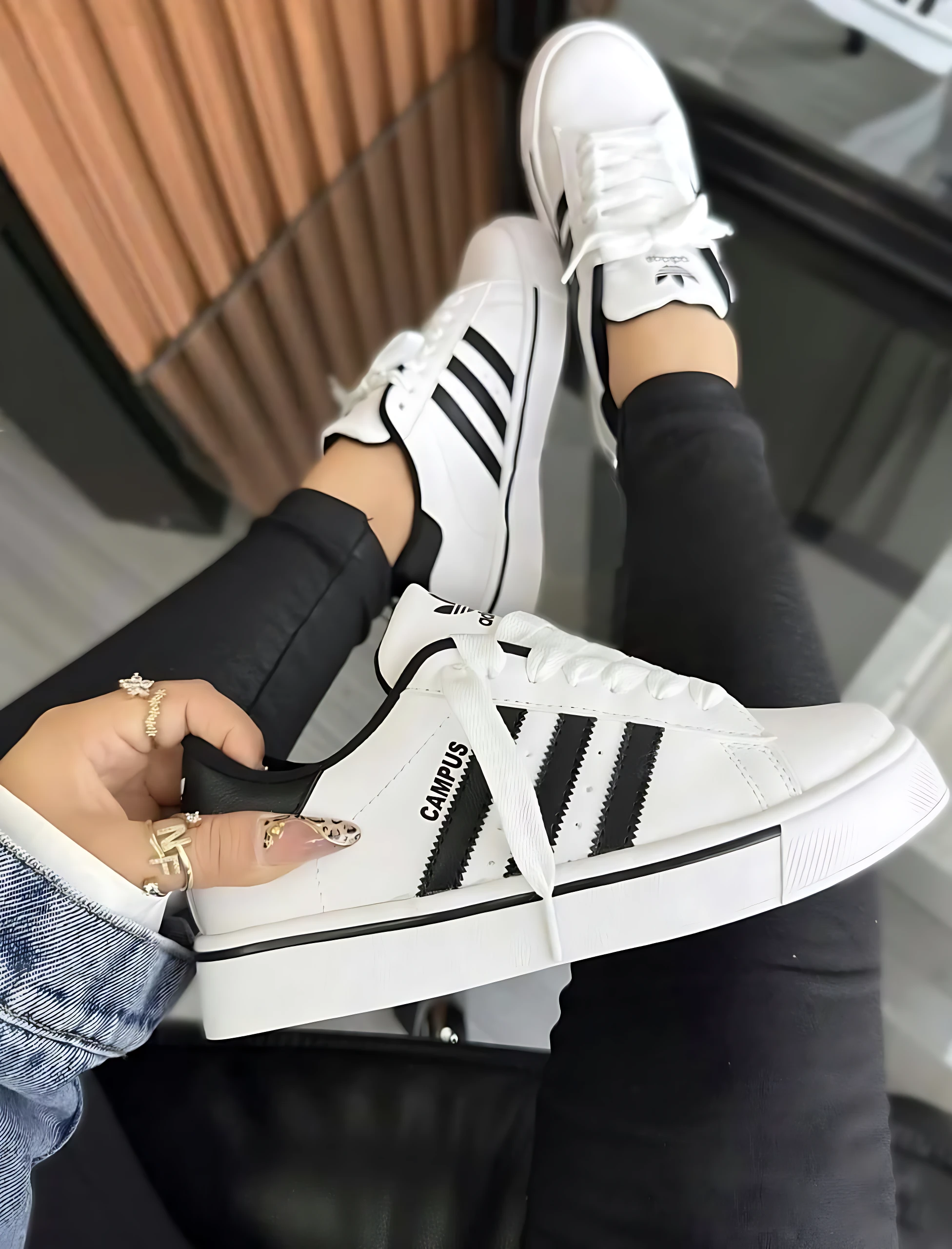 Zapatilla Adidas Campus blanca con tres rayas negras, suela acolchada y cordones blancos, vista desde arriba con pie derecho levantado y mano sosteniendo el lateral izquierdo.