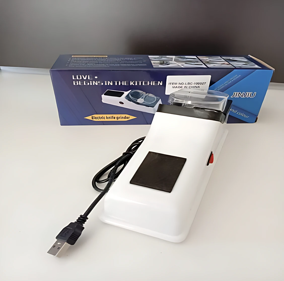 Afilador eléctrico automático blanco con cable USB, base rectangular y caja azul al fondo que muestra el producto en uso.