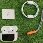 Vista superior de Airpods ANC tercera generación en estuche blanco abierto, con cable de carga enrollado y manual, sobre fondo de césped artificial.