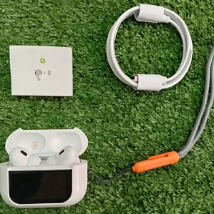 Vista superior de Airpods ANC tercera generación en estuche blanco abierto, con cable de carga enrollado y manual, sobre fondo de césped artificial.