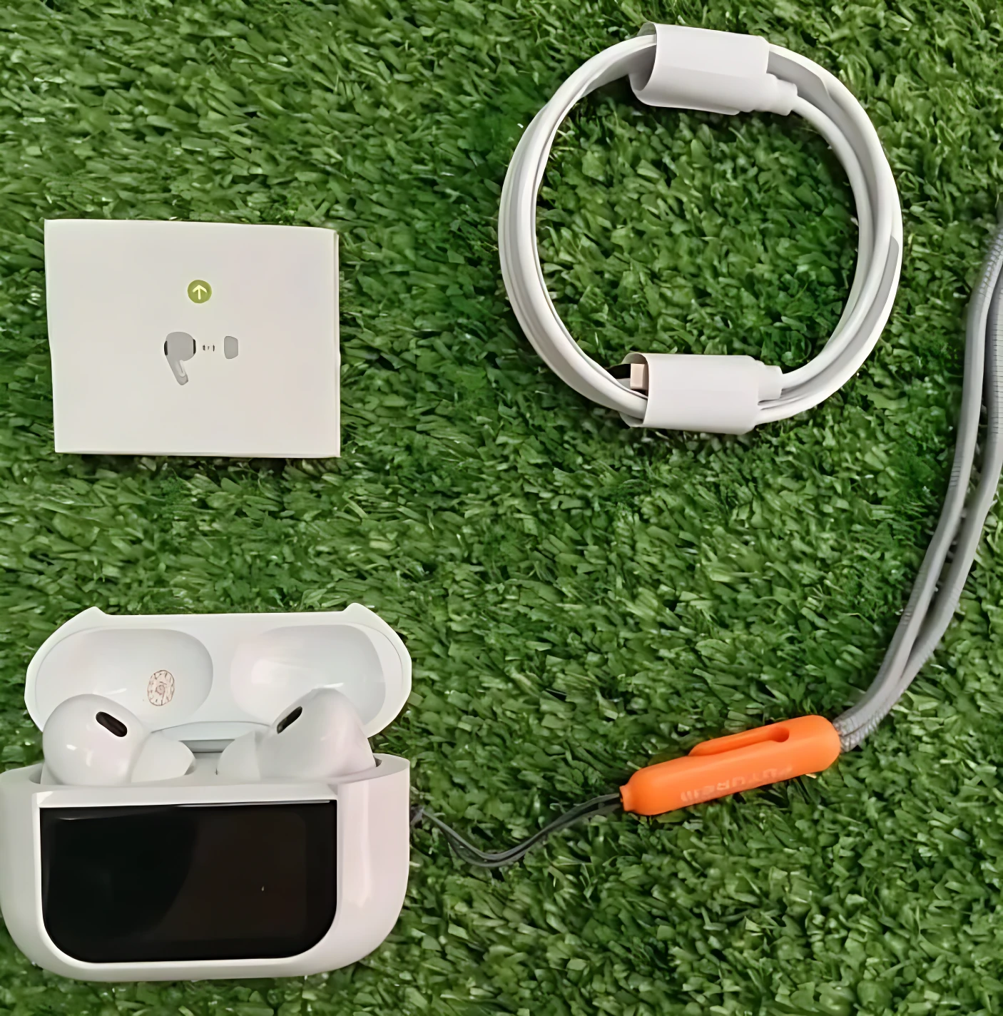 Vista superior de Airpods ANC tercera generación en estuche blanco abierto, con cable de carga enrollado y manual, sobre fondo de césped artificial.