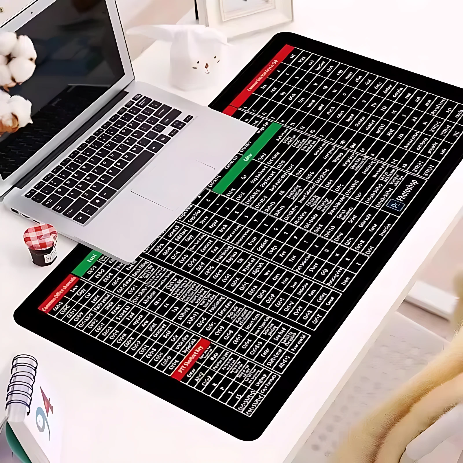 Alfombrilla de escritorio XXL negra con atajos visuales de software, colocada sobre mesa blanca junto a laptop plateada y accesorios de oficina.