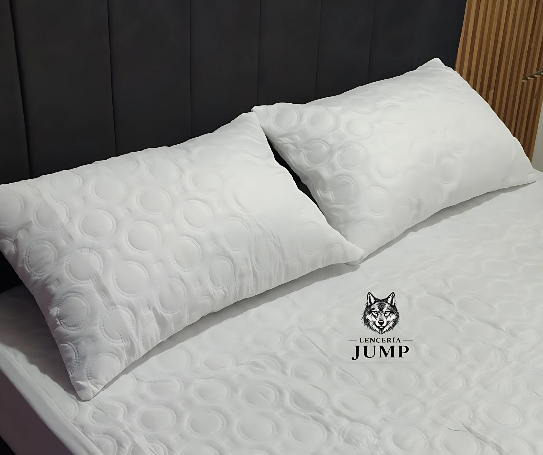 Dos almohadas blancas con textura de círculos acolchados, sobre cama con cabecera oscura. Modelo termosellado, relleno de algodón siliconado.
