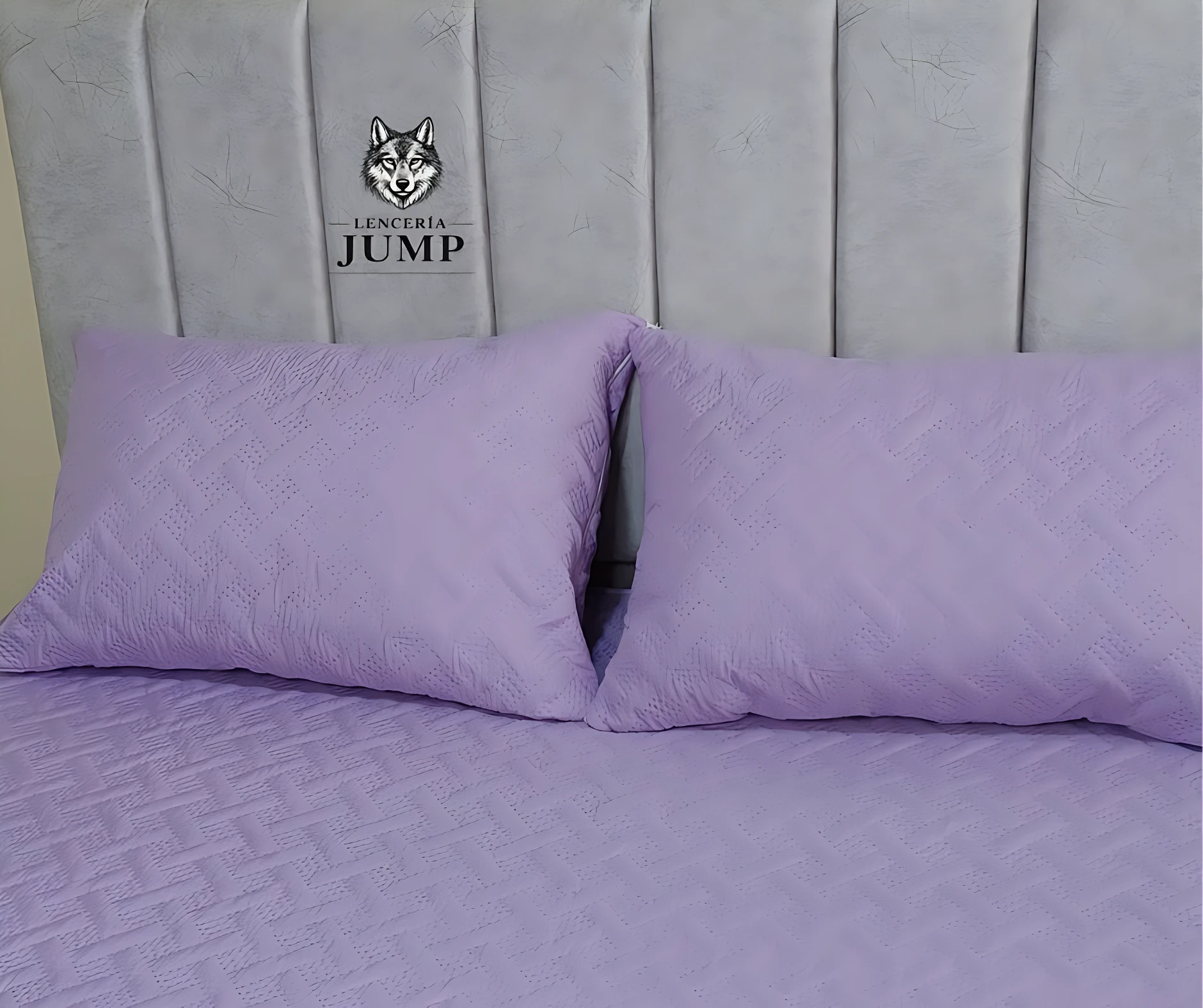 Set de dos fundas de almohada en tono lila con textura geométrica, sobre cama con cabecero acolchado gris claro y logo visible.