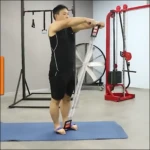 Hombre usando bandas de tracción con agarres metálicos, en posición de estiramiento frontal sobre colchoneta azul, en gimnasio con equipo rojo al fondo.
