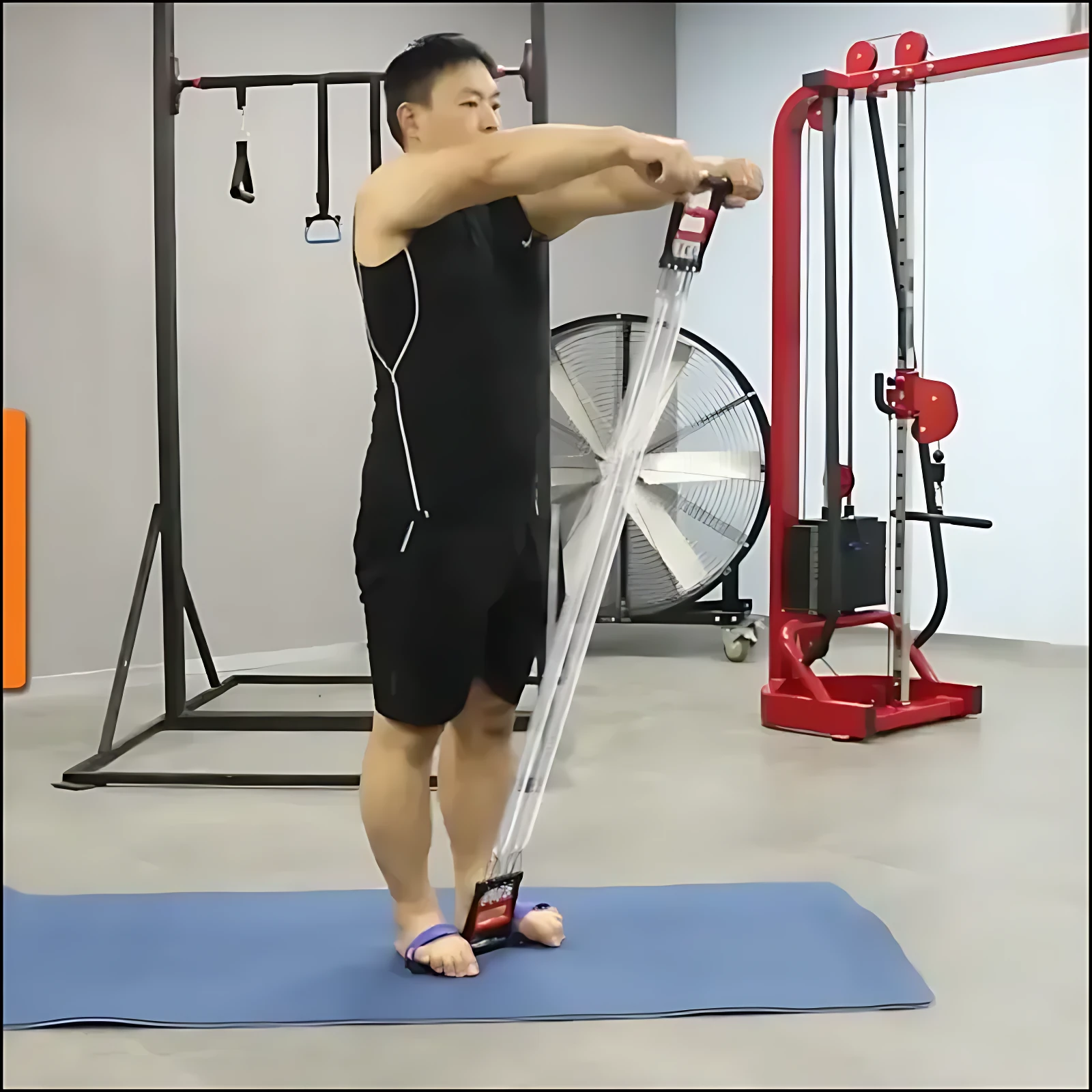 Hombre usando bandas de tracción con agarres metálicos, en posición de estiramiento frontal sobre colchoneta azul, en gimnasio con equipo rojo al fondo.