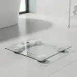 Báscula de vidrio templado cuadrada con patas blancas y pantalla LCD, sobre piso de cerámica en baño moderno.