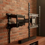 Base TV de pared en color negro, con brazo articulado y soporte metálico, montada sobre estante de madera frente a pared de ladrillo visto.