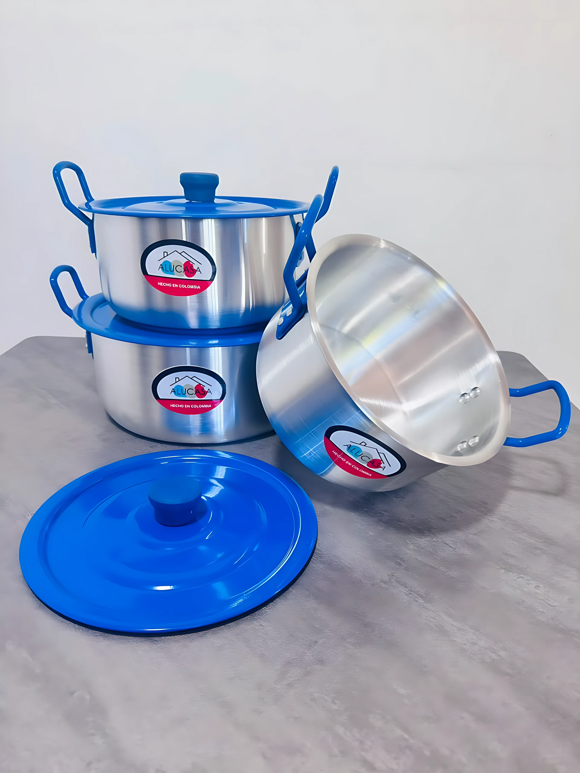 Batería de ollas de acero inoxidable con asas azules y tapas, mostrando tres recipientes apilados y uno inclinado sobre superficie gris.