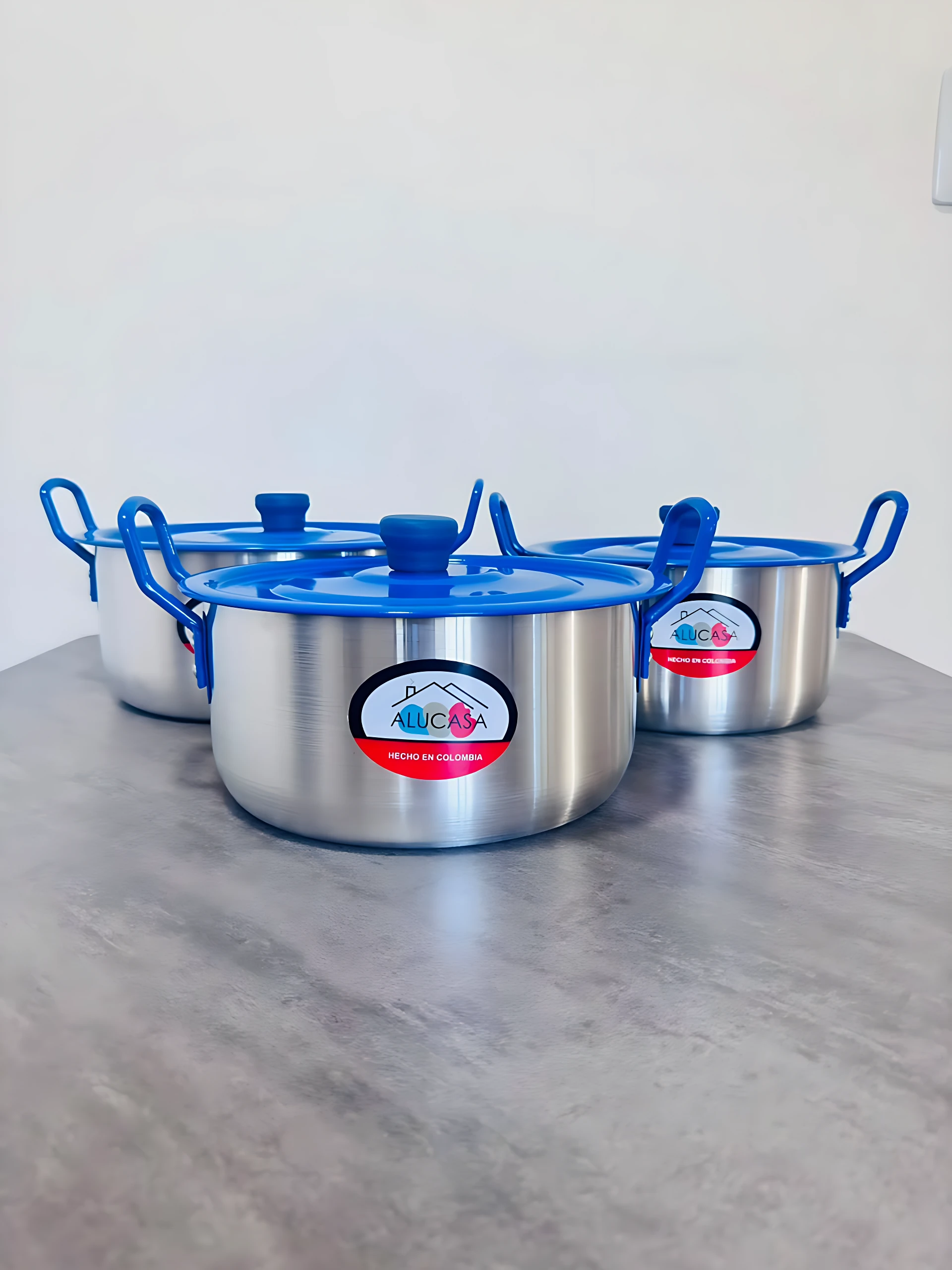 Set de tres ollas de acero inoxidable con tapas y asas azules, dispuestas en fila sobre superficie gris.
