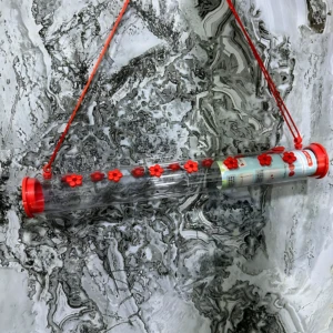 Bebedero para colibríes con tapas rojas antiparasitos, colgado por cuerdas rojas sobre fondo de mármol blanco y gris.