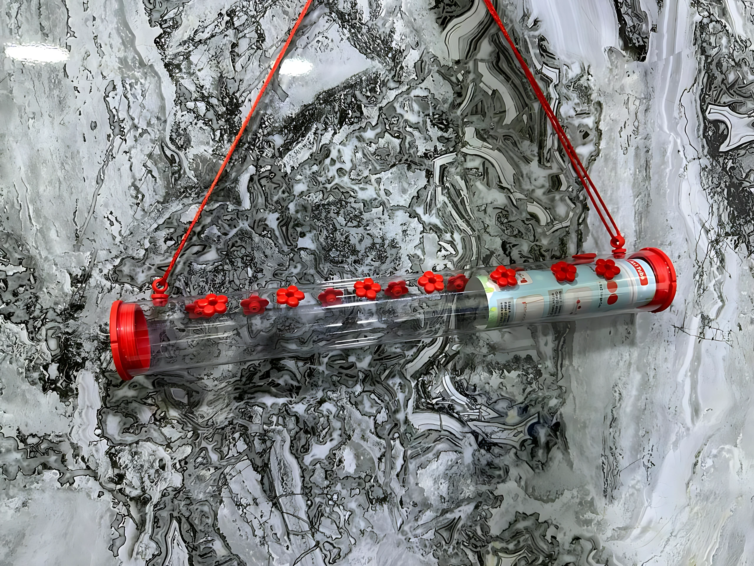 Bebedero para colibríes con tapas rojas antiparasitos, colgado de cuerdas rojas sobre fondo de mármol blanco y gris.