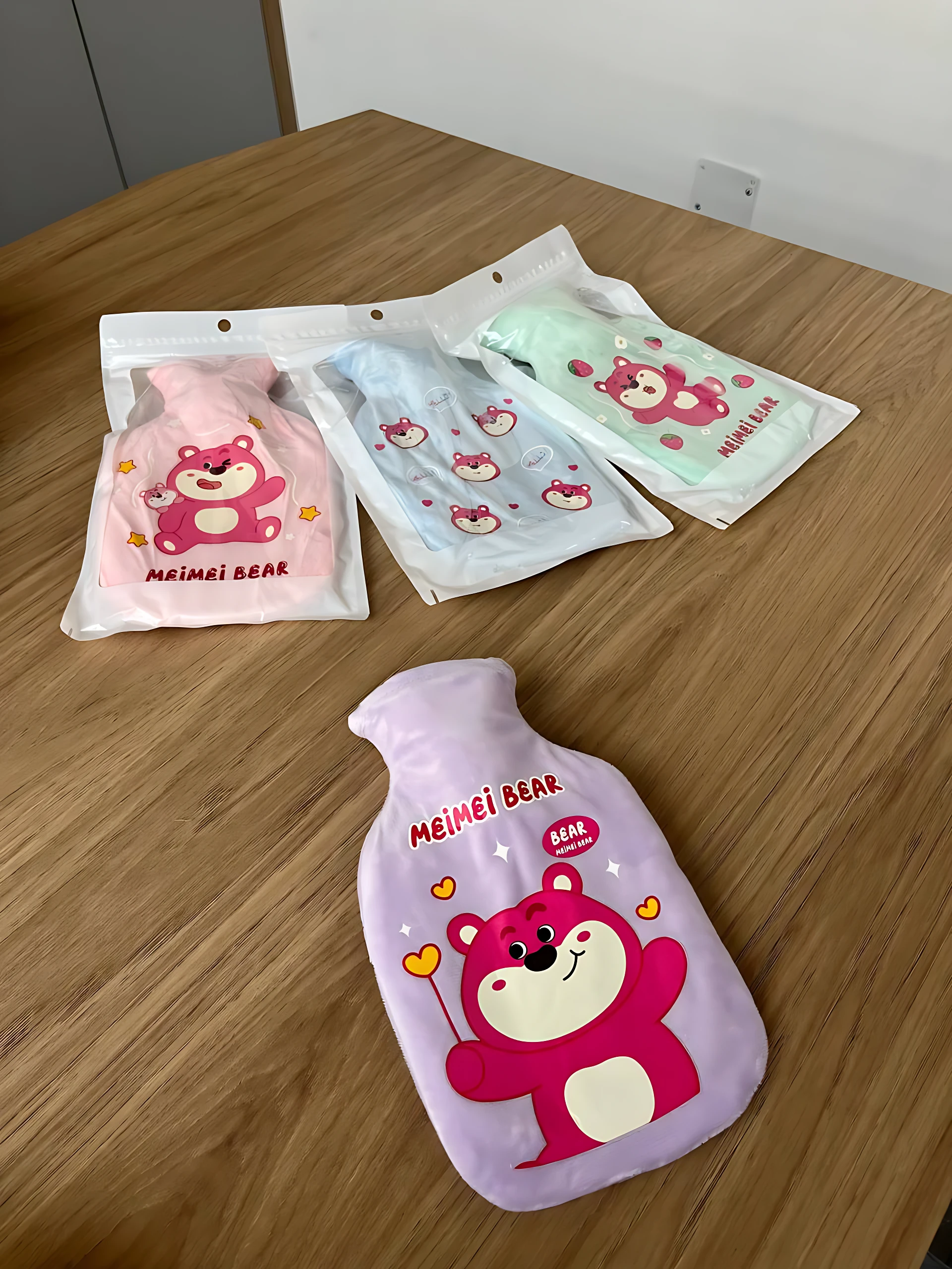 Bolsa de agua caliente Meimei Bear en color lila con oso rosa, mostrando variante en tres colores dentro de su empaque individual.