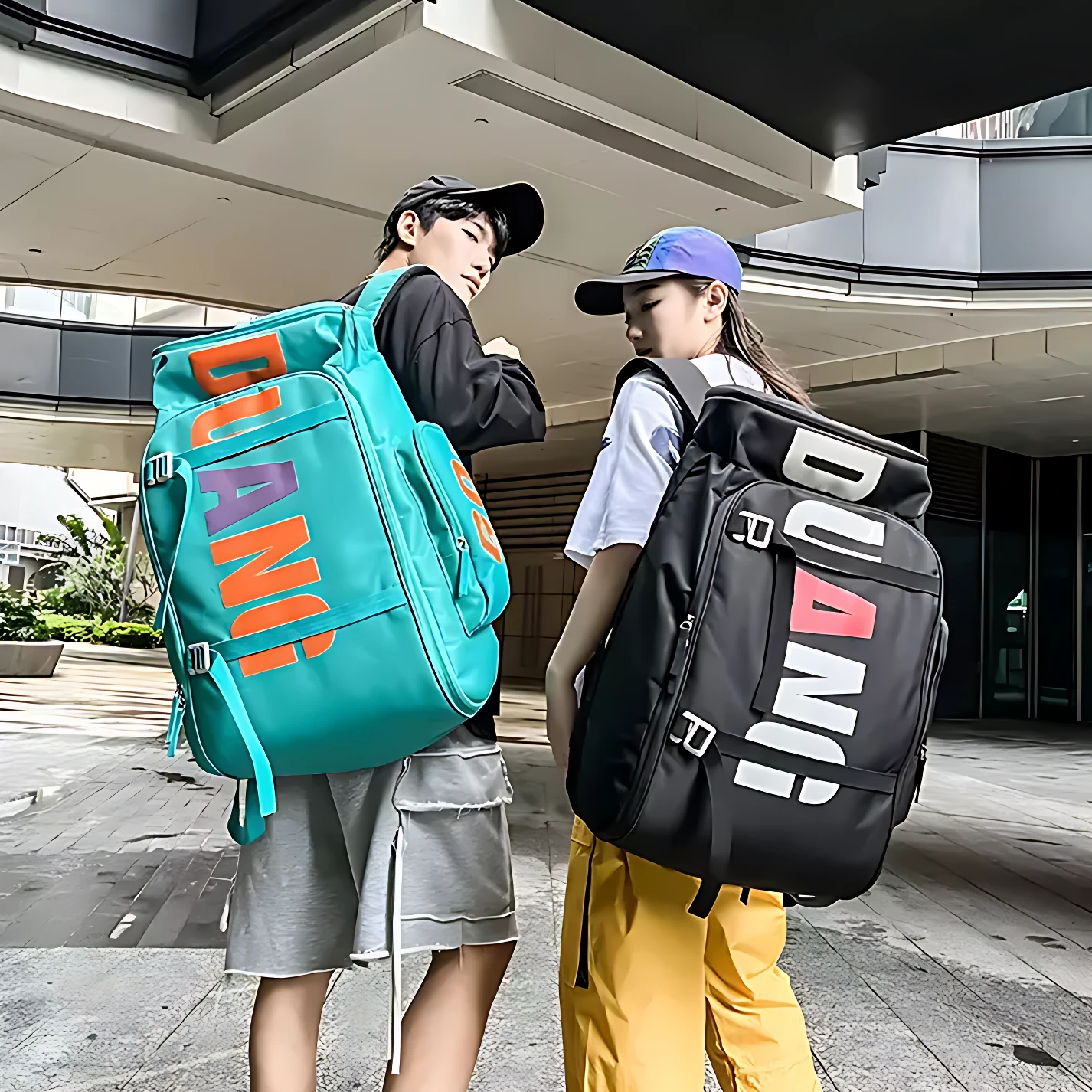 Dos modelos usan mochilas grandes con diseño urbano; una en color turquesa con letras naranjas y moradas, la otra negra con letras blancas y rojas, ambas con múltiples compartimentos visibles.