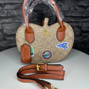 Bolso Coach monograma color beige con asas de cuero marrón, correa ajustable y parches deportivos visibles.