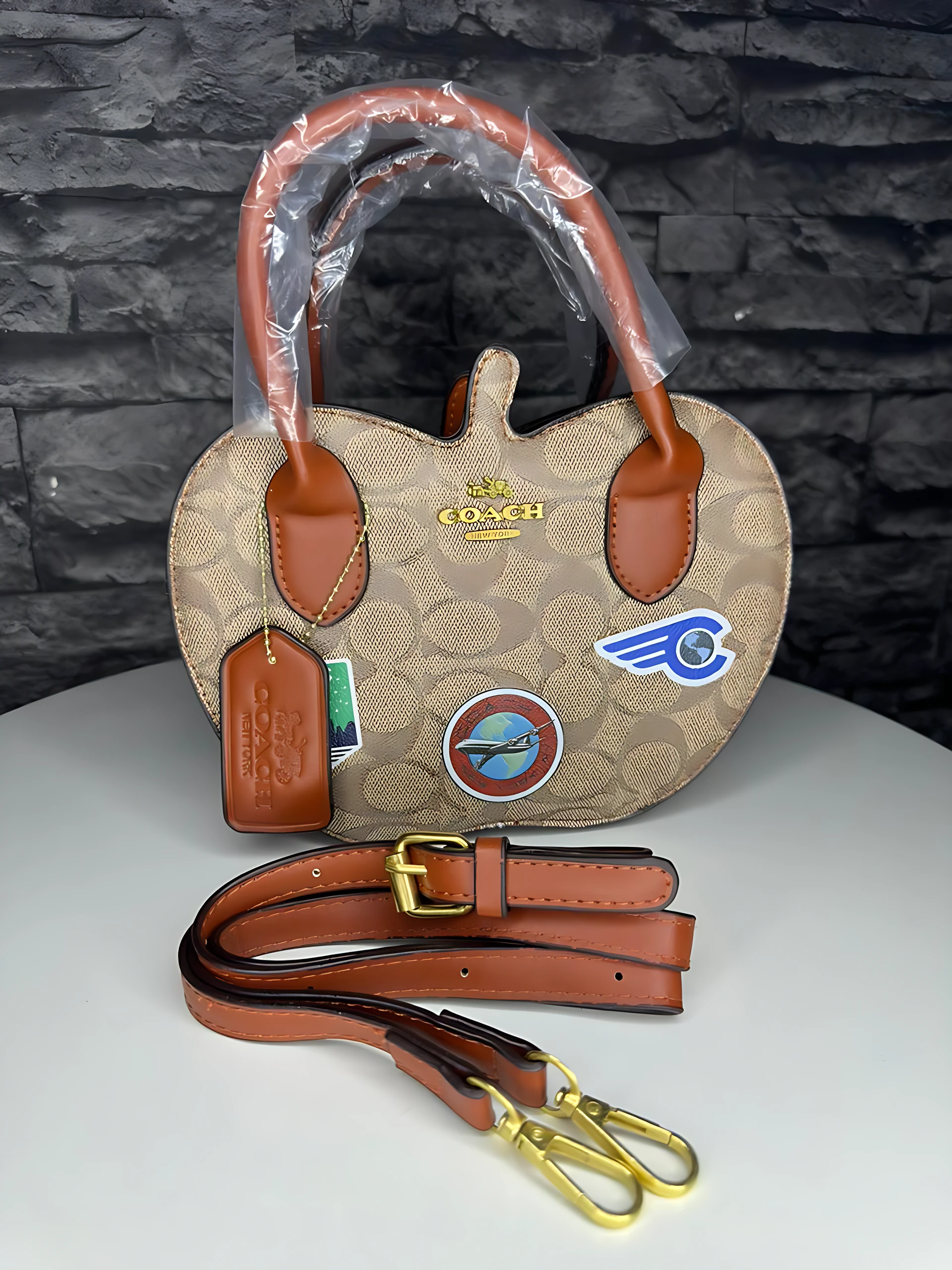 Bolso Coach monograma color beige con asas de cuero marrón, correa ajustable y parches deportivos visibles.