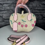 Bolso Coach monograma color crema con estampado de cerezas rosas y correa ajustable en color rosa, sobre mesa blanca.