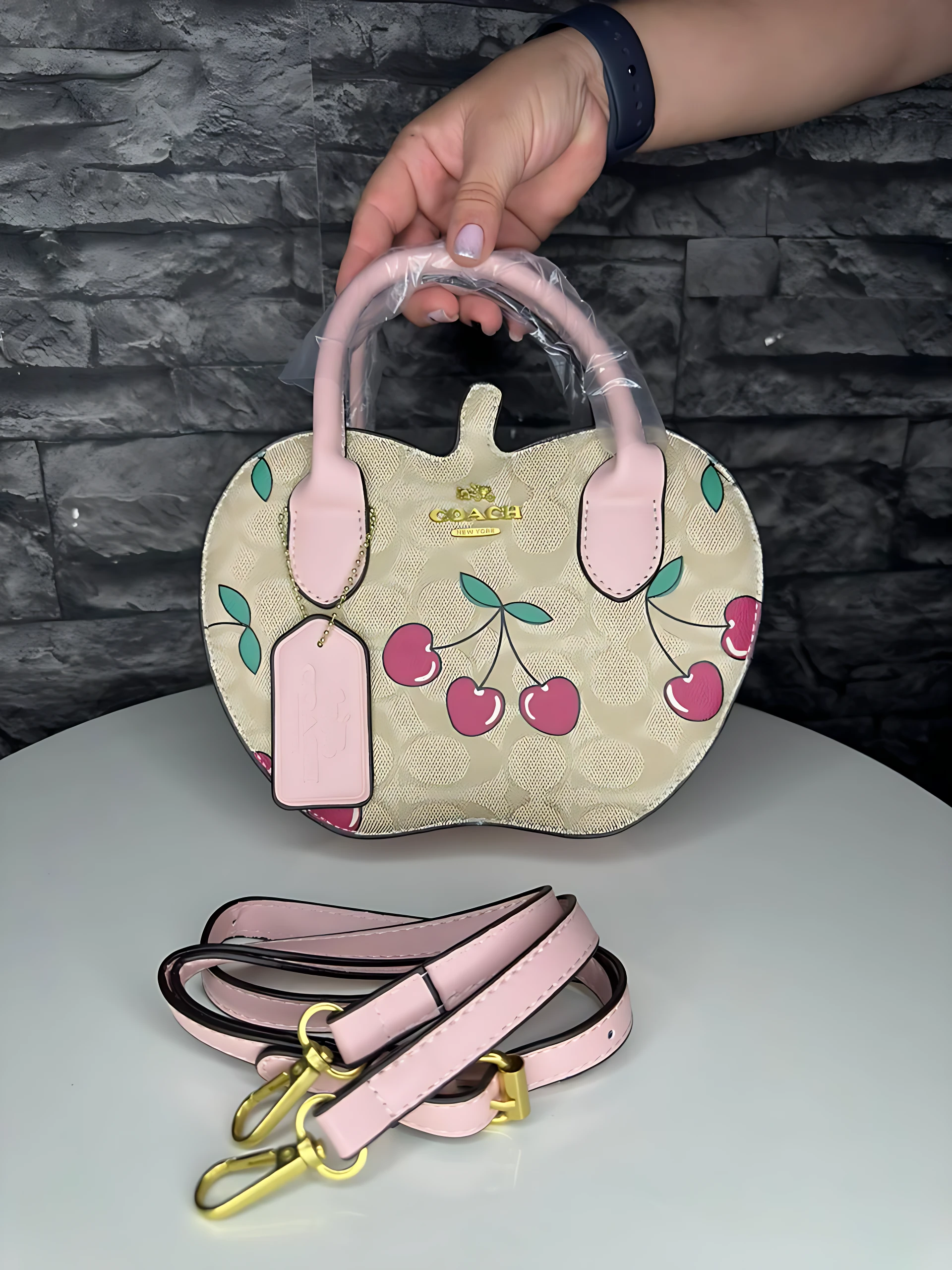 Bolso Coach monograma color crema con estampado de cerezas rosas y correa ajustable en color rosa, sobre mesa blanca.