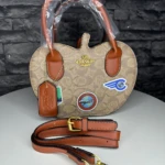 Bolso Coach monograma color beige con asas y correa ajustable marrón, detalles en cuero y parches deportivos visibles.