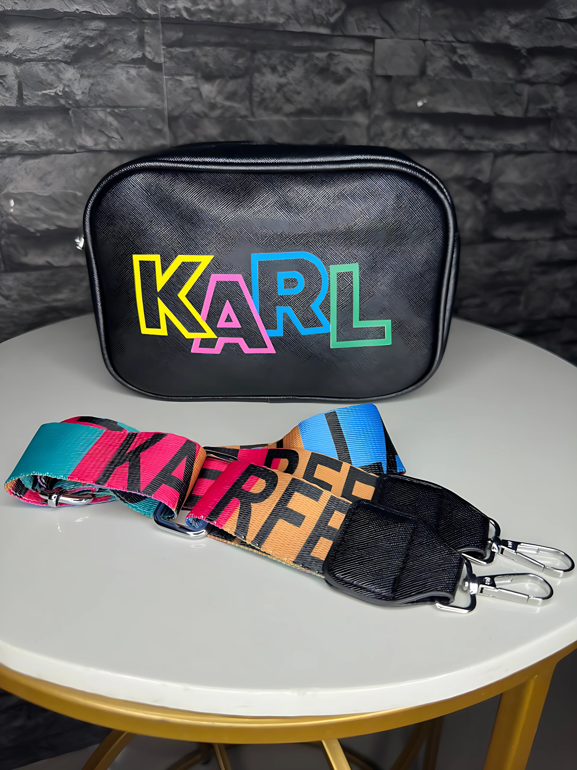 Bolso crossbody negro con logo KARL en colores neón, acompañado de correa ajustable multicolor con hebilla metálica, sobre mesa redonda blanca frente a pared de ladrillo gris.