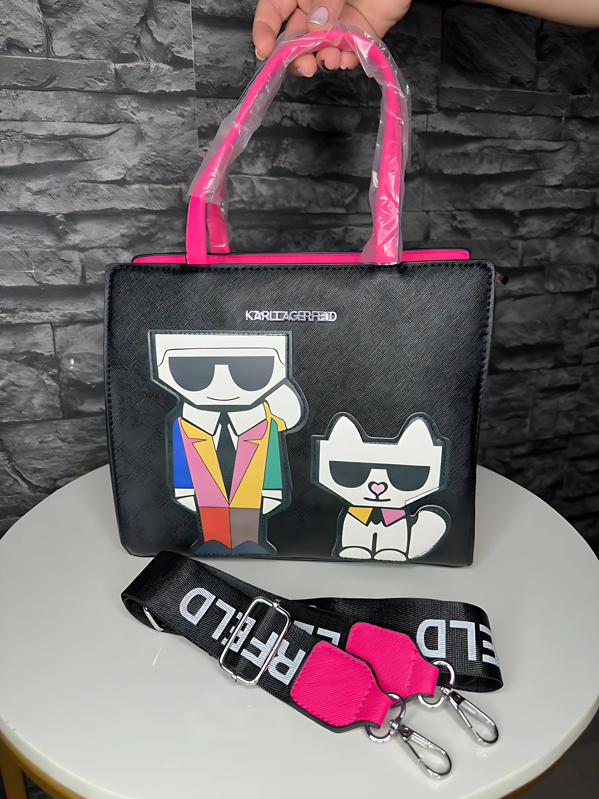 Bolso de mano negro con asas rosas y diseño de Karl Lagerfeld y su gato, sobre mesa blanca con correa desmontable incluida.