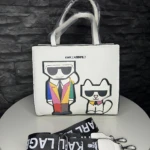 Bolso de mano blanco KARL LAGERFELD con ilustraciones de Karl y su gato, sobre mesa redonda; incluye correa negra con logo.