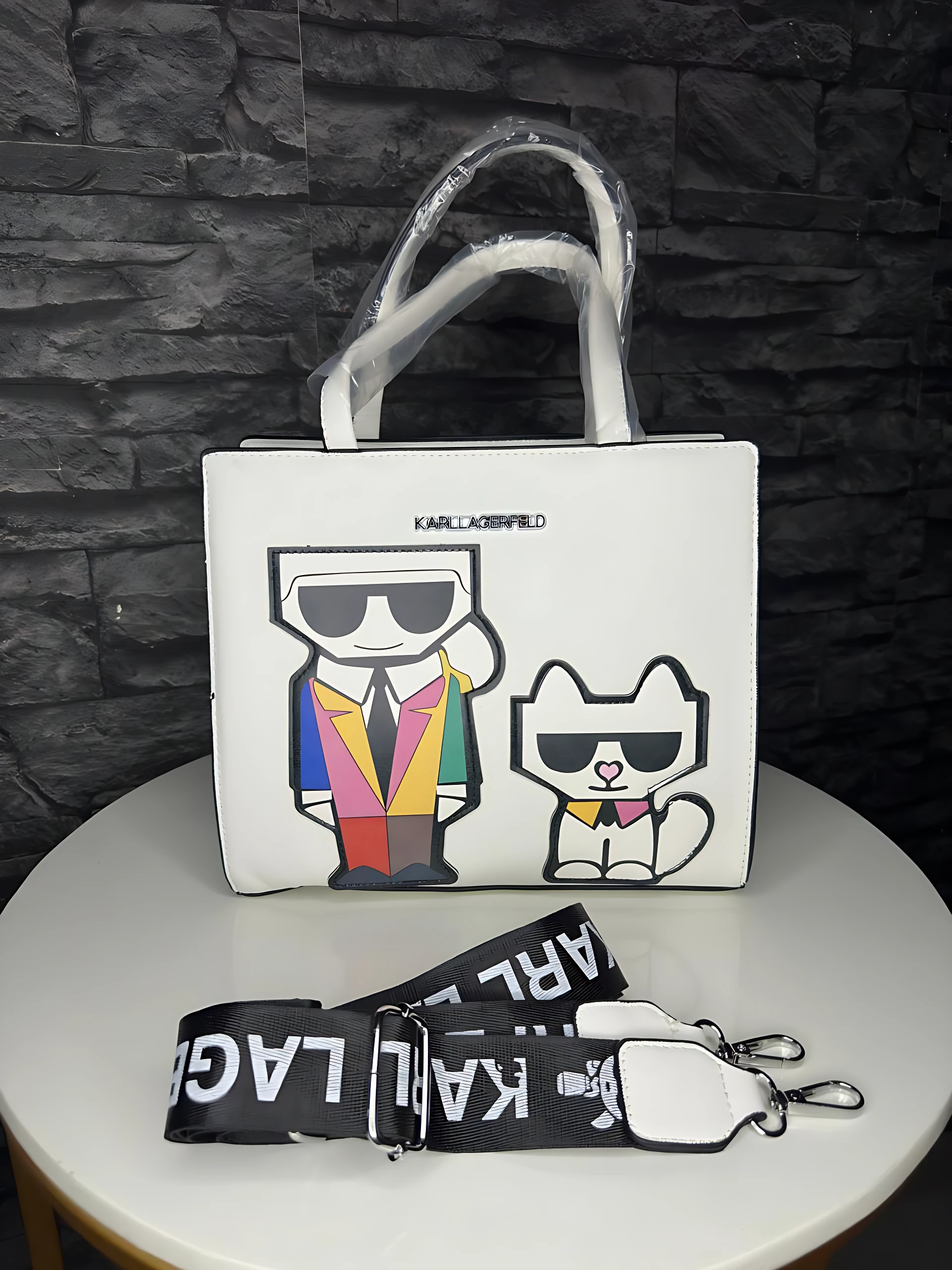 Bolso de mano blanco KARL LAGERFELD con ilustraciones de Karl y su gato, sobre mesa redonda; incluye correa negra con logo.