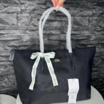 Bolso tote negro Lacoste con asas blancas y lazo decorativo, sostenido por una mano frente a pared de piedra.