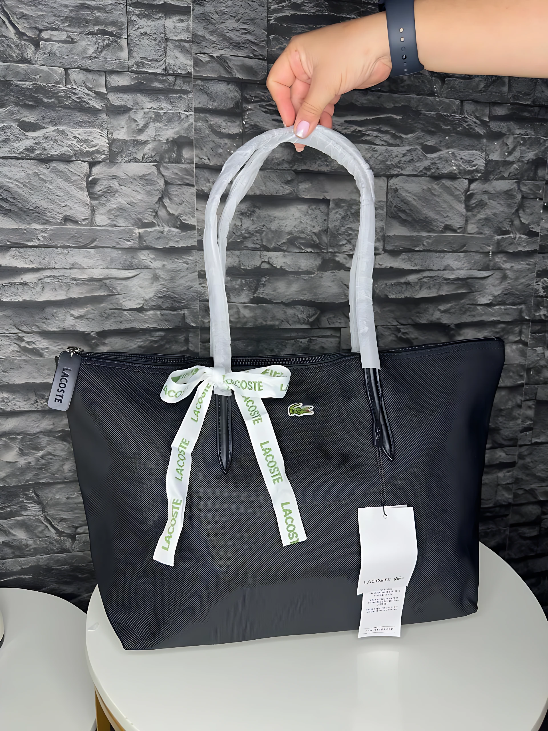 Bolso tote negro Lacoste con asas blancas y lazo decorativo, sostenido por una mano frente a pared de piedra.
