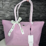 Bolso tote Lacoste en tono lila claro, con asas de mismo color y lazo decorativo con cinta blanca que lleva la marca. Incluye etiqueta colgante con logo de la marca.