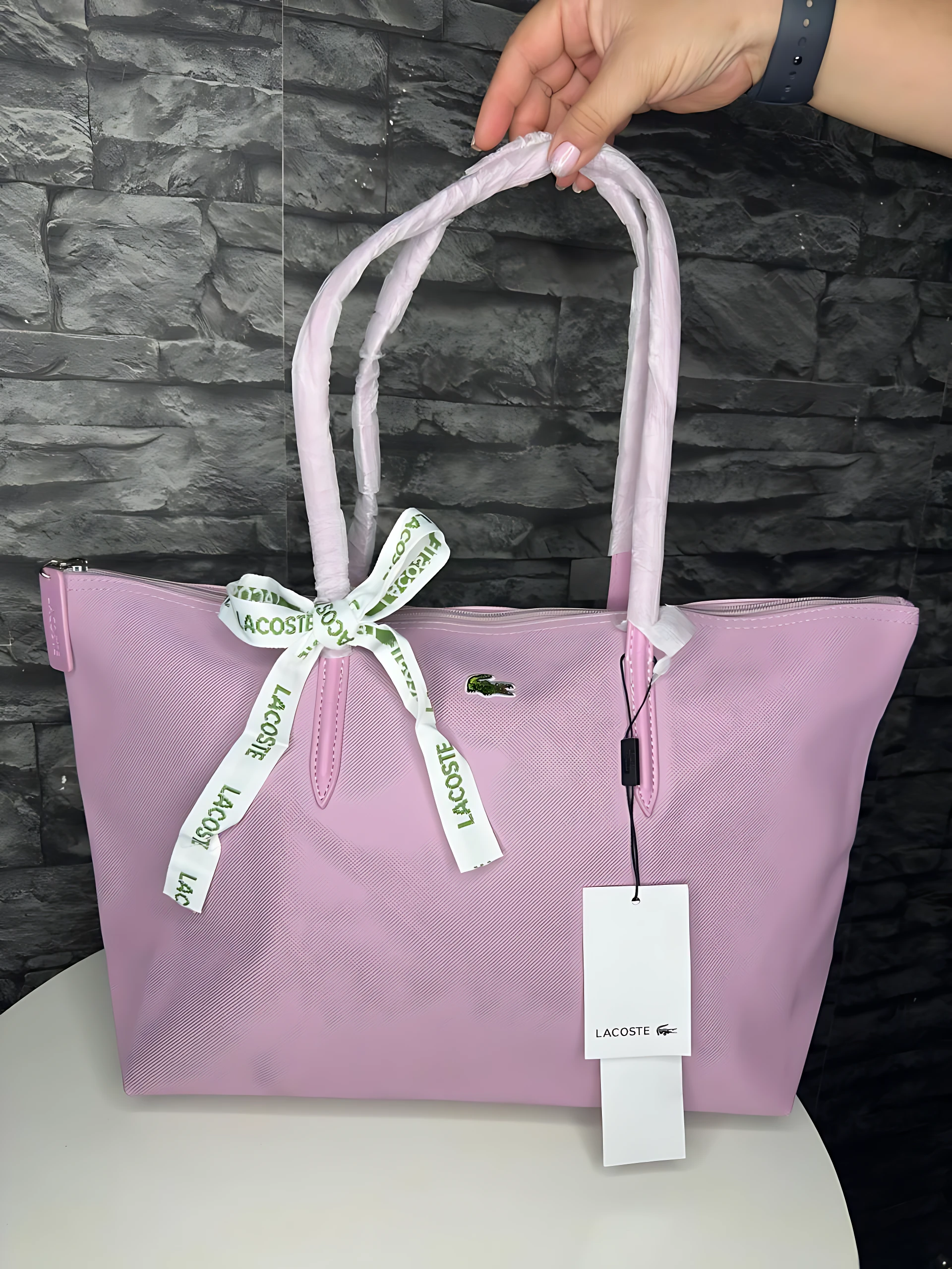 Bolso tote Lacoste en tono lila claro, con asas de mismo color y lazo decorativo con cinta blanca que lleva la marca. Incluye etiqueta colgante con logo de la marca.
