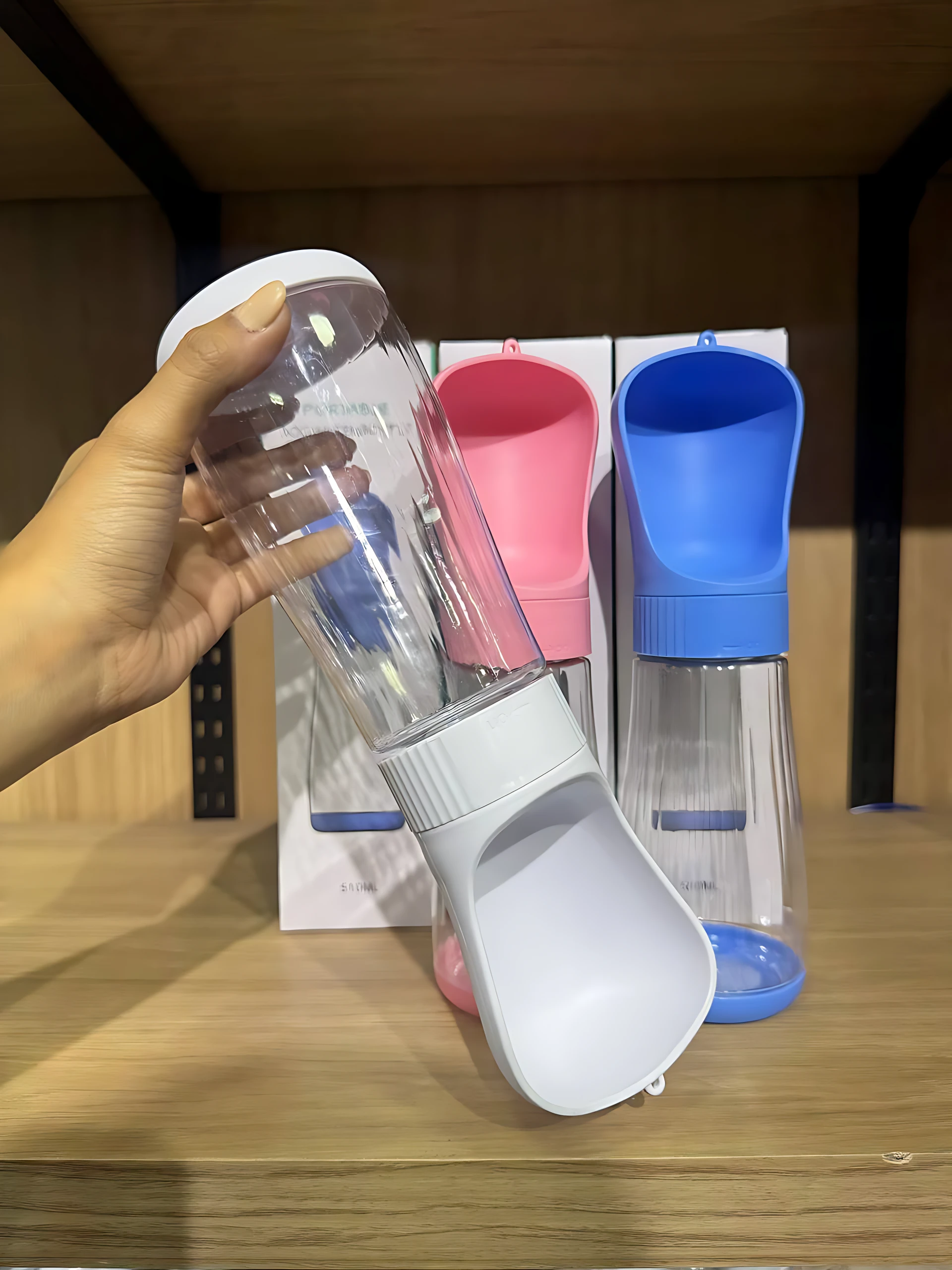 Botella portatil para mascotas con bebedero integrado, en color blanco, siendo sostenida por una mano. Al fondo se observan dos unidades adicionales, una de color rosa y otra de color azul, ambas sobre sus respectivas cajas de presentación.