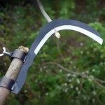 Cabezal de hoz en acero al manganeso montado en mango de madera, con hoja curva negra y borde blanco, listo para cortar césped y ramas.