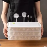 Caja de almacenamiento para congelador con tapa blanca y tres niveles transparentes, mostrando empaquetados de alimentos congelados, sostenida por persona con delantal negro.