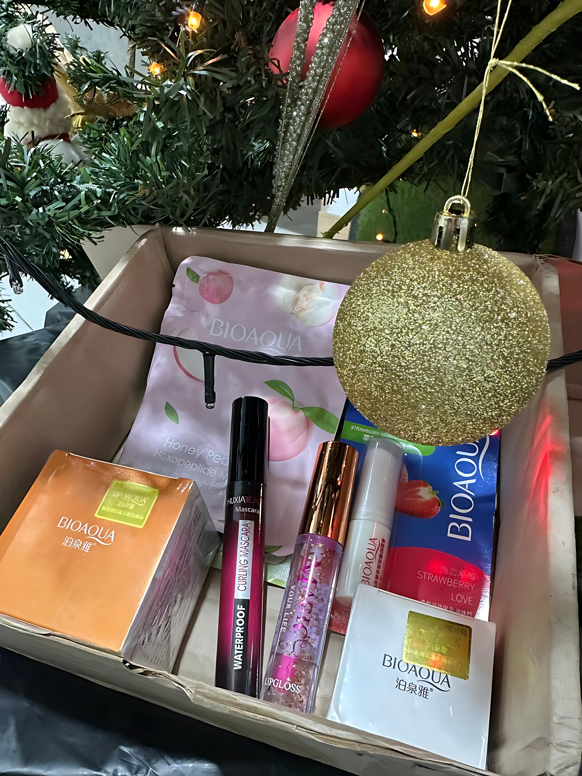 Caja de regalo con productos de cuidado facial y maquillaje de Bioaqua, incluyendo mascarilla, labial, brillo de labios y crema, dispuestos sobre fondo navideño con árbol y bola dorada.