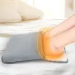 Calentador de pies eléctrico portátil en tela suave gris, con efecto de calor visual en zona de tobillo, sobre cama blanca con almohadas.