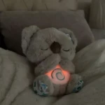 Peluche koala de peluche gris con luz cálida en el pecho y patas estampadas, sentado sobre manta suave en cama.