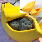 Cama en forma de banano amarilla con dos gatos gris dentro, mostrando interior acolchado y diseño de fruta realista.