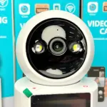 Cámara de videovigilancia blanca con pantalla integrada, visión nocturna y dos luces LED frontales, sobre base con botón de encendido.