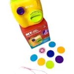 Cámara de juguete amarilla con detalles en morado, diseñada para niños, junto a su caja roja y accesorios incluidos: lentes de colores, proyector y catapulta. Ideal para motricidad fina y creatividad.