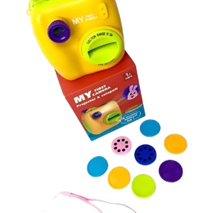 Cámara de juguete amarilla con detalles en morado, diseñada para niños, junto a su caja roja y accesorios incluidos: lentes de colores, proyector y catapulta. Ideal para motricidad fina y creatividad.