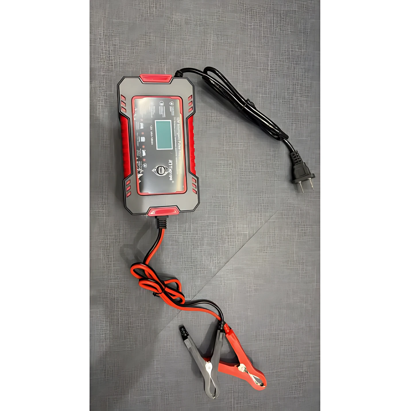 Cargador inteligente de batería 12V con pantalla digital, cables rojo y negro y pinzas de conexión, sobre superficie gris texturizada.