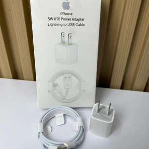 Pack de cargador iPhone 5W con cable Lightning, incluye adaptador y cable enrollado, sobre superficie blanca con fondo de madera.