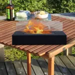 Chimenea de etanol portátil negra con flama realista sobre mesa de madera al aire libre, acompañada de botella y plato.