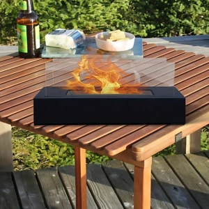 Chimenea de etanol portátil negra con flama realista sobre mesa de madera al aire libre, acompañada de botella y plato.