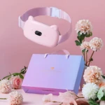 Cinturón masajeador abdominal recargable en color rosa pastel con diseño de orejas de gato, mostrando panel de control y banda elástica. Incluye caja de regalo con asa y flores decorativas alrededor.