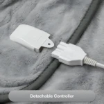 Control desmontable blanco de cobija eléctrica sobre tela de felpa gris, con cable conectado y botón visible.