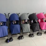 Seis sillas de paseo tipo sombrilla en colores azul, gris, negro y rosa, dispuestas en fila sobre suelo blanco con ruedas y capotas visibles.